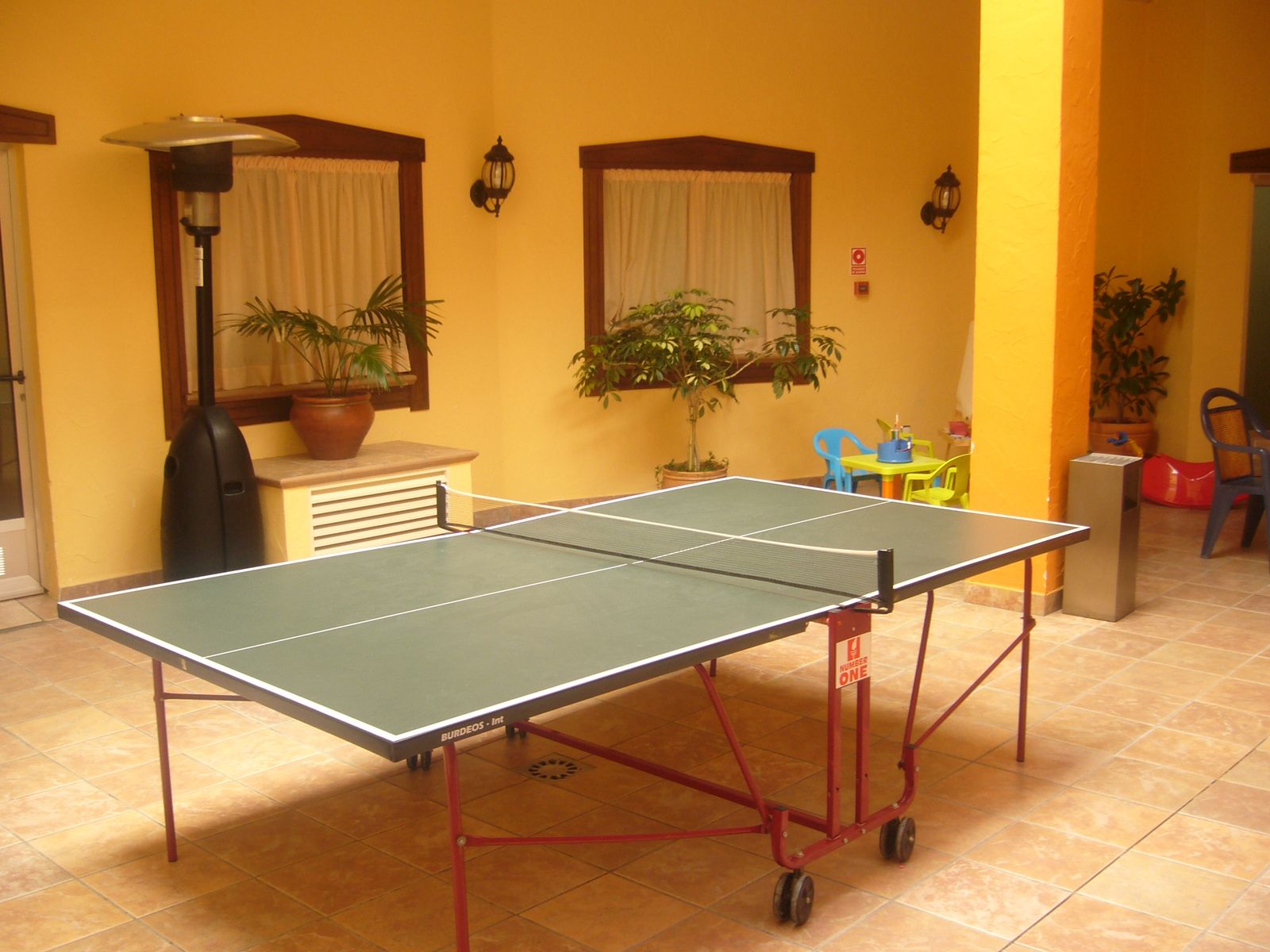 Green table tennis table with its net up in a tiled common area with yellow walls at Hotel Traíña